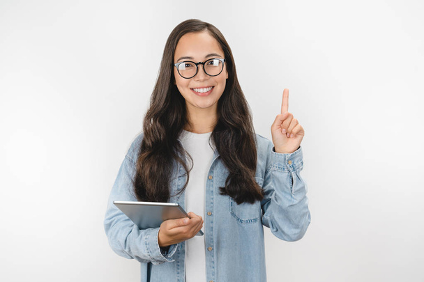 Excited asian woman holding digital tablet and pointing finger up at copy space while standing isolated over white background - Фото, изображение