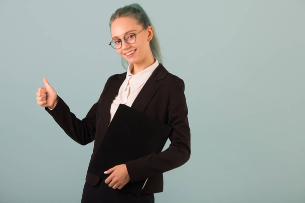 beautiful young woman in glasses and a black suit on a blue background with a folder in her hands - Фото, зображення