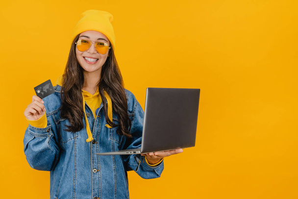 Happy woman in hat holding credit card and laptop isolated over yellow background - Fotó, kép