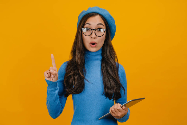 Asian woman holding digital tablet and pointing finger up at copy space while standing isolated over yellow background - Foto, Bild