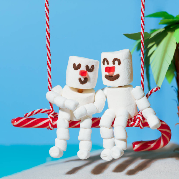 Zwei Liebhaber von Marshmallow-Schneemännern sitzen auf einer Süßigkeitenschaukel. Weihnachtsurlaub am Strand. - Foto, Bild