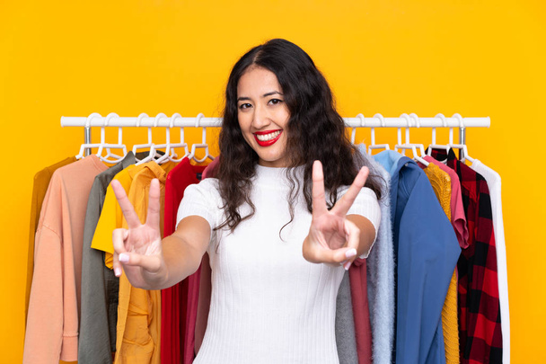 Mujer de raza mixta en una tienda de ropa sonriendo y mostrando señal de victoria
 - Foto, Imagen