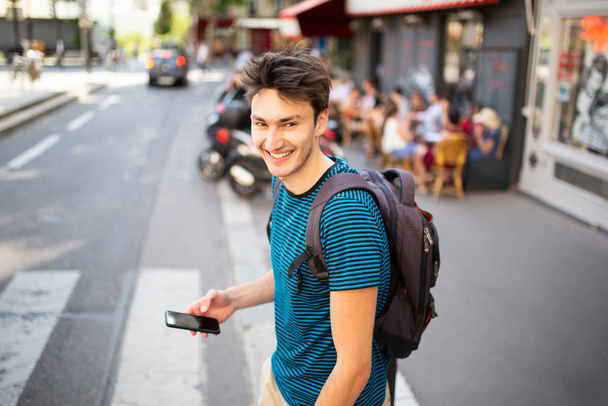 Πορτρέτο χαμογελαστός νεαρός άνδρας περπάτημα με τσάντα και κινητό τηλέφωνο στο δρόμο της πόλης - Φωτογραφία, εικόνα