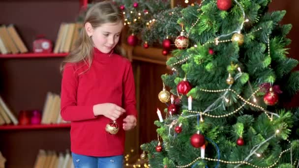 Cute girl in a red sweater decorates the Christmas tree in the room with decorative balls. Preparing for Christmas and New Year. Holidays - Video