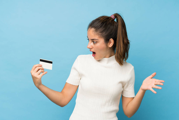 Young brunette girl over isolated blue background holding a credit card - Fotografie, Obrázek