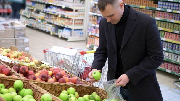 Молодой человек в плаще собирает яблоки на прилавке в супермаркете
 - Фото, изображение