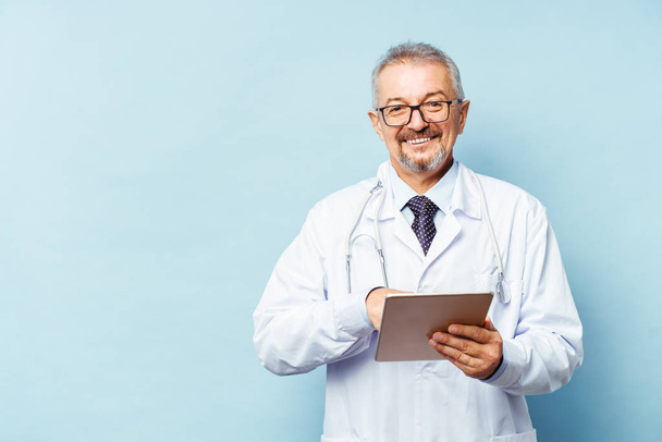 Medical senior doctor with a stethoscope. On a blue background. A medic holds a tablet in his hands and makes an appointment with the clinic. Disease prevention concept - 写真・画像
