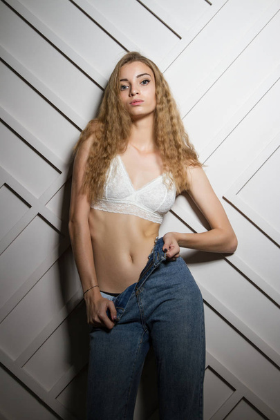 Beautiful young girl in bra and jeans stands leaning against the wall - 写真・画像