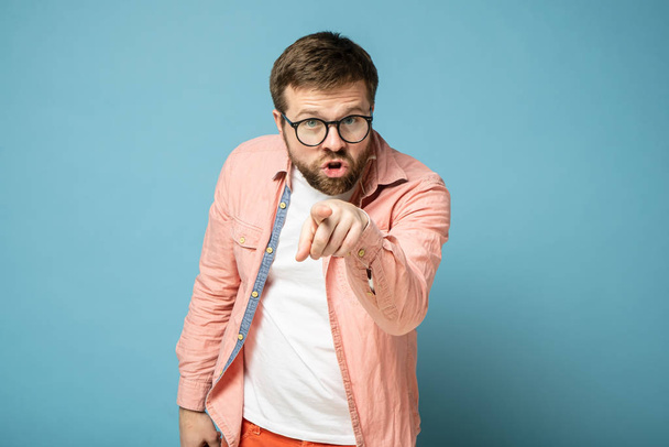 Agressieve man met een bril wijst met een wijsvinger naar iemand en geeft de schuld, kijkend naar de camera. Geïsoleerd op een blauwe achtergrond. - Foto, afbeelding