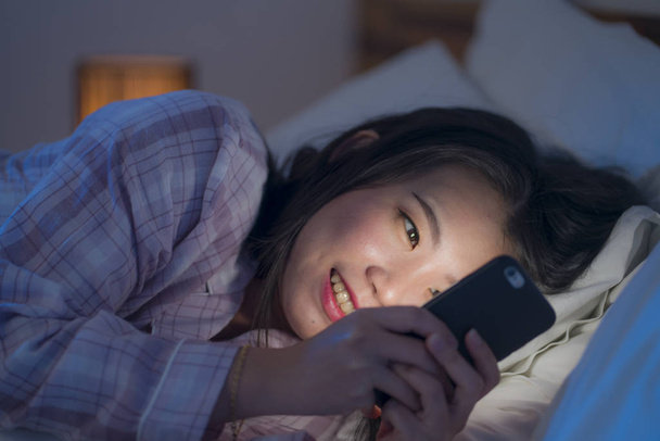 young beautiful and happy sweet Asian Korean woman with in pajamas enjoying with mobile phone app in bed at night in internet social media addiction concept - Photo, image
