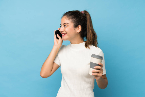jong brunette meisje over geïsoleerde blauwe achtergrond holding koffie weg te nemen en een mobiele - Foto, afbeelding