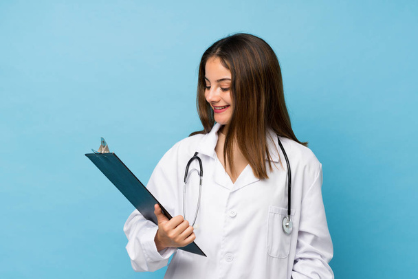 Young brunette girl over isolated blue background with doctor gown and holding a folder - Foto, imagen