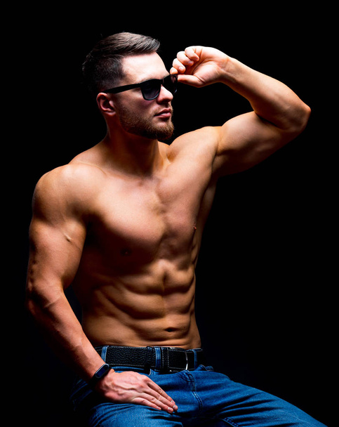 Well build handsome man without a shirt posing in studio. Strong body and cool face. Male wearing glasses, sitting on a chair with hands on legs. selective focus. Closeup. - Fotografie, Obrázek