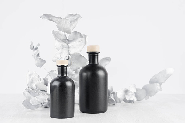 Blank cosmetic glass black bottles with beige bungs decorated silver leaves on light white wood table as mock up for advertising, branding identity, design. - 写真・画像