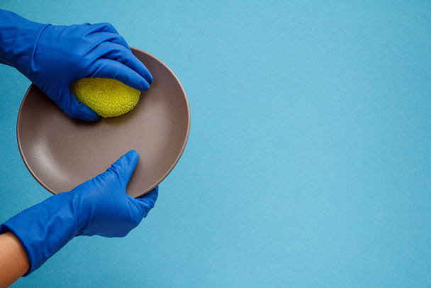 Women's hands in protective gloves with sponge and plate on blue background. - Fotoğraf, Görsel
