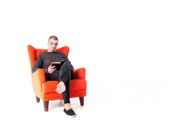 Un joven atractivo está sentado en un sillón rojo y leyendo un libro. Aislado sobre un fondo blanco. Copiar espacio
. - Foto, imagen