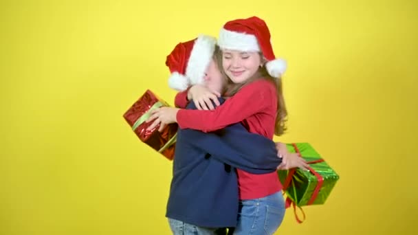 Cute children in santa claus hats give presents to each other and hug, on a yellow studio background. Holidays and fun. Christmas and New Year. - Video