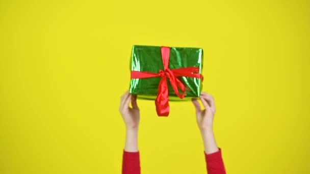 Children's hands raise up and hold a gift, a large gift box wrapped in green paper with a red ribbon on a studio yellow background - Felvétel, videó