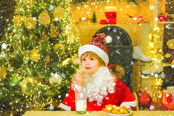 Kerstman met kerstkoekjes en melk tegen kerstboom achtergrond. Kind in de sneeuw. Koekjes voor kinderen Kerstman. Kid Santa Claus genieten in geserveerd peperkoek cake en melk. - Foto, afbeelding