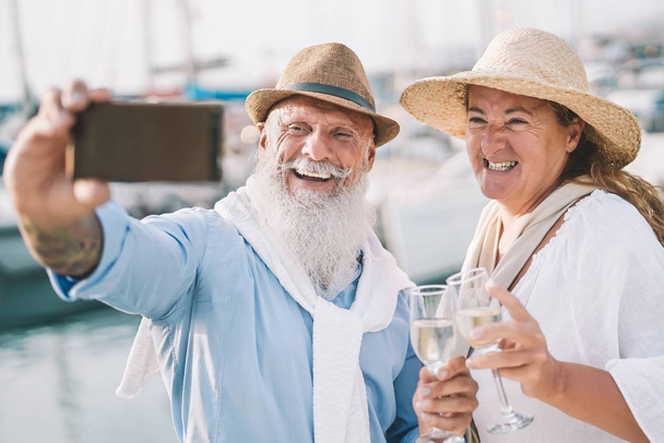 Seniors couple taking selfie with mobile smartphone on sailboat vacation - Happy people having fun celebrate drinking champagne on boat trip - Relationship love and romantic travel dating concept - Photo, Image