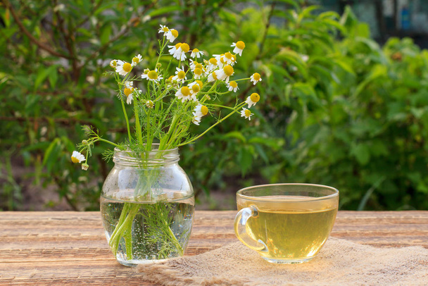 Glazen kopje groene thee met witte kamillebloemen in een glazen pot buiten. - Foto, afbeelding