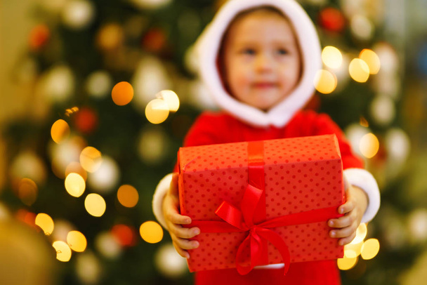 Christmas gift in the hands of a child. Hands of little boy in santa costume with Christmas present. Smiling funny kid holding red gift box on the background of lights. Holidays, New year concept. - Photo, Image