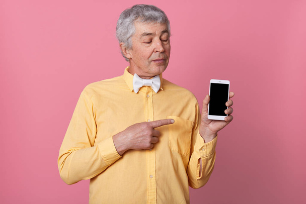 Close up portrait of senior man wearing yellow shirt with white bowtie showing black blank smart phone screen, looking at his device, posing over pink background, looks serious and concentrated. - 写真・画像