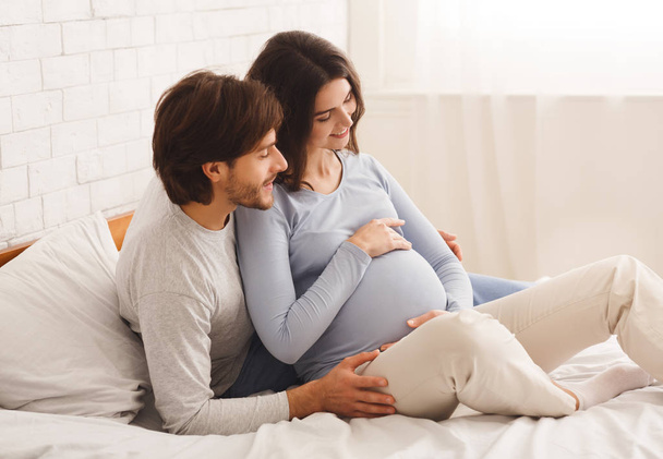 Boldog várandós pár ül az ágyon otthon, élvezi a jövőbeli szülőség - Fotó, kép