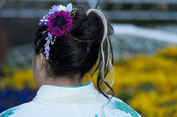 Beautiful Japanese teenager back wearing traditional kimono with close-up on hair ornament celebrating the Coming of Age Day in Fuji City, Japan. Background with defocused yellow flowers. - Photo, Image
