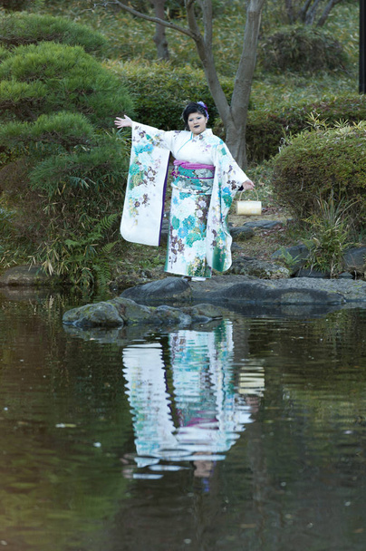 Красивый японский подросток, одетый в традиционное кимоно, стоит с распростертыми руками и празднует День совершеннолетия в Фудзи, Япония. Фон с зелеными кустами листвы
.  - Фото, изображение