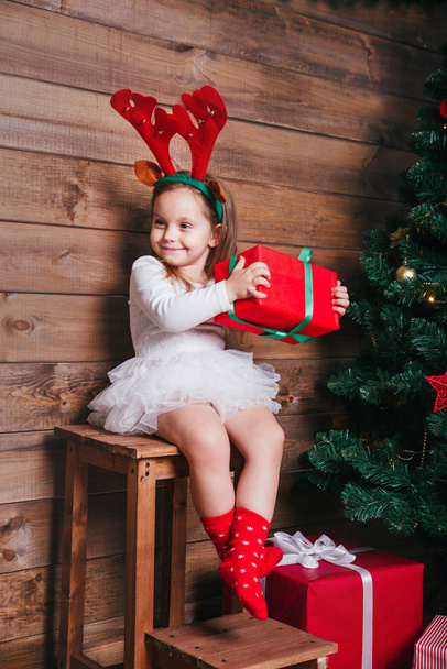 Smiling funny child in red deer horns holding Christmas gift in hand near Christmas tree at home - Foto, immagini