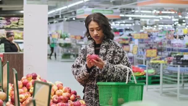 Woman buying apples in supermarket. Medium of african american female choosing fruits in store and places it into shopping cart. Woman take red apple in marketplace. Vegan sales concept in 4K, UHD - Πλάνα, βίντεο