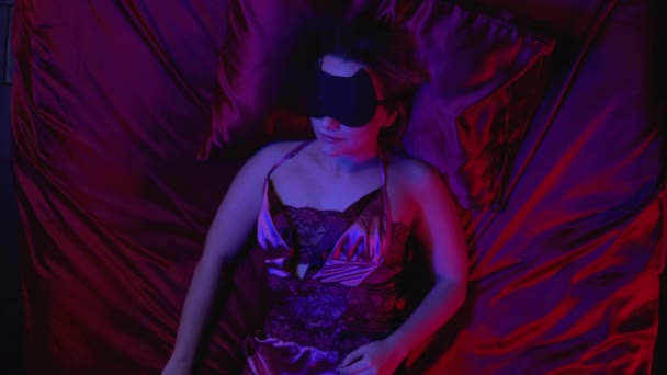 Beautiful young woman sleeping on silk bed linen alone, sleep mask on face - Materiał filmowy, wideo