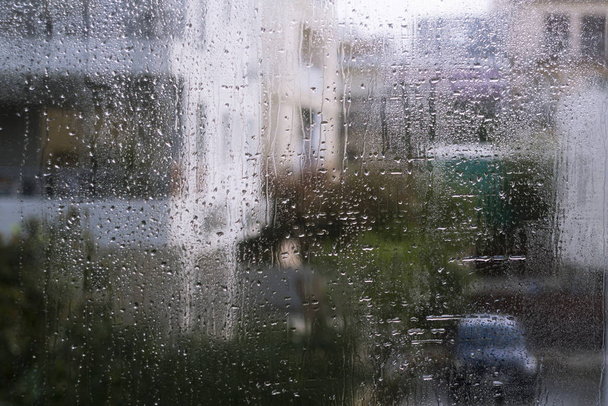 Мокрий віконний келих з великою кількістю крапель дощу і видом на розмиті силуети будинків, дерев і автомобіля на вулиці
 - Фото, зображення