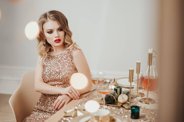 young beautiful woman in gold luxury dress. hollywood hairstyle and red lips. New year table setting. Ceramic plate with fork and knife on a sequin napkin. The concept of a festive dinner. - Photo, Image