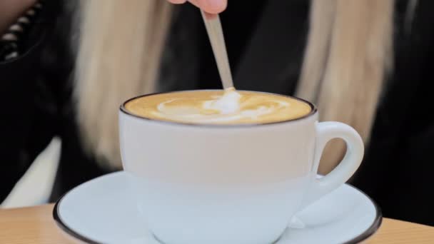 Une femme blonde caucasienne non reconnue agite du sucre avec une petite cuillère dans une tasse en céramique de café cappuccino
.  - Séquence, vidéo