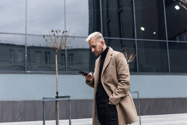 Businessman  looking at smartphone while standing in beige coat near building. - Fotografie, Obrázek