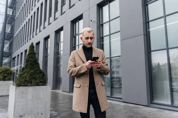 Young Attractive Businessman Standing near Big Modern Office Building. Typing a Message on his Smartphone. - Foto, immagini