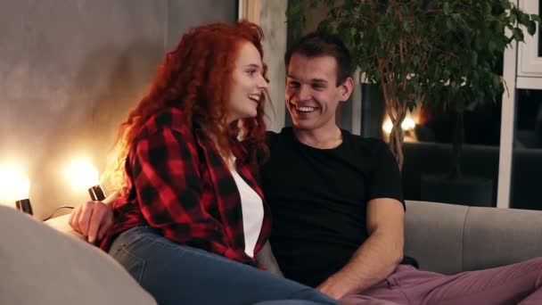 Close up footage of loving, positive man and happy young curly longhaired woman embracing and chatting while relaxing on sofa in their home, stylish living room with house plant on the background - Filmati, video