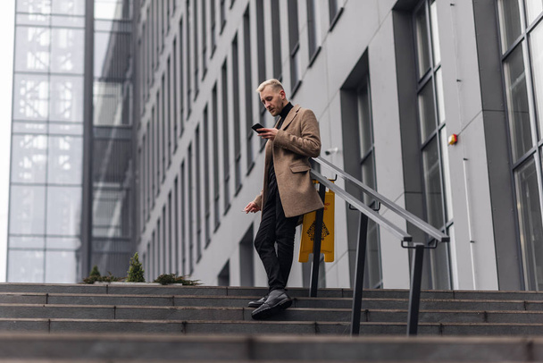 Handsome businessman standing in a beige coat with crossed legs and looking at the phone. - Foto, afbeelding