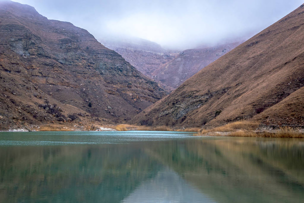 A small lake is surrounded by desert mountains without trees and vegetation. Mountains are reflected in turquoise water. The tops of the mountains are covered with fog, which gives the picture a mystical look. - Photo, Image