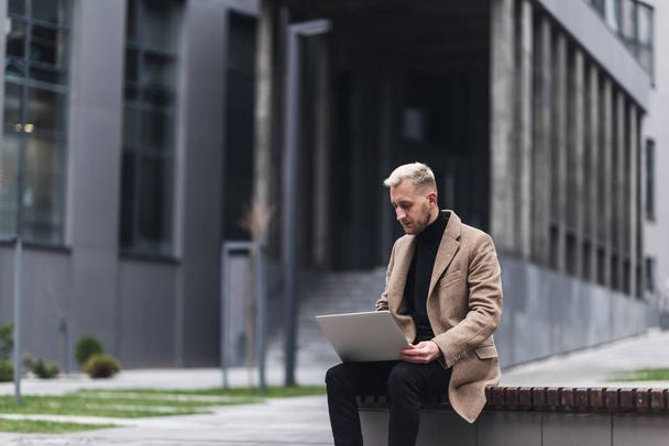 Stylish man sitting on the streets of the city near modern office centre and working with his laptop. - Foto, Bild