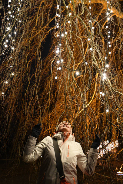 Young woman under hanging branches of huge willow tree decorated with shiny Christmas lights garlands glowing in night darkness. Fairy tale illuminations of tree branches. Holiday scene - Photo, Image
