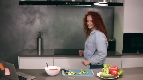 Happy active red headed young woman holding spoon as a microphone singing songs and dancing while cooking in kitchen, carefree girl preparing vegetable salad healthy food having fun alone at home - Imágenes, Vídeo