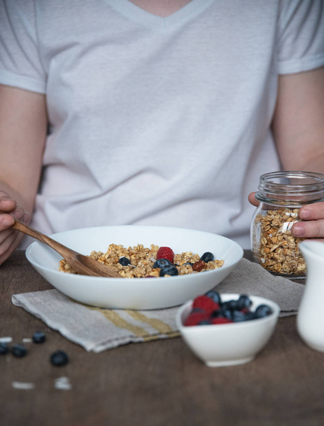 a young girl pours muesli into a white plate for Breakfast with berries at a brown wooden table. Vegan food - Photo, Image
