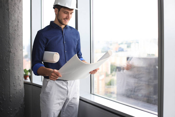 Confident young business man in hardhat holding blueprint and looking away while standing indoors. - Fotoğraf, Görsel