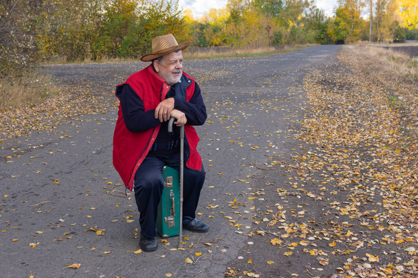 Starší turista s vycházkovou holí sedí na starém zeleném kufru na silnici, zatímco čeká na autobus na Ukrajině - Fotografie, Obrázek