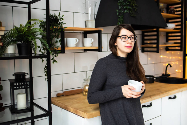 Mladá žena v tmavě šedých šatech v moderní kuchyni, v kuchyni pije čaj. - Fotografie, Obrázek