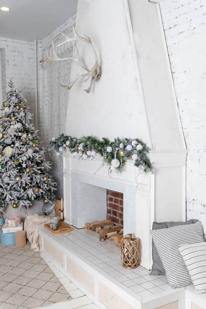 リビングルームで暖炉とクリスマスの装飾。白い暖炉のクリスマスガーランド。白の装飾、金の玉、クリスマスツリー、新年のインテリア。祭りの家の装飾。 - 写真・画像