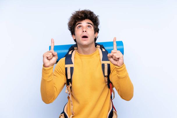 Jovem montanhista homem com uma grande mochila sobre fundo azul isolado apontando com o dedo indicador uma ótima ideia - Foto, Imagem